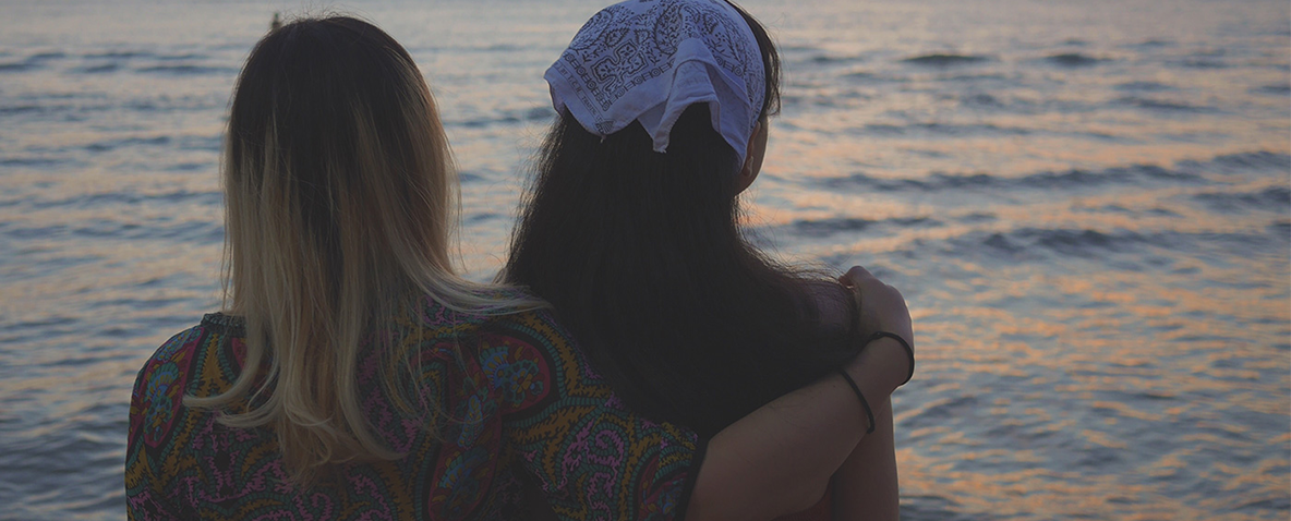 Two women sitting along the water with arms around each other's shoulder at sunset