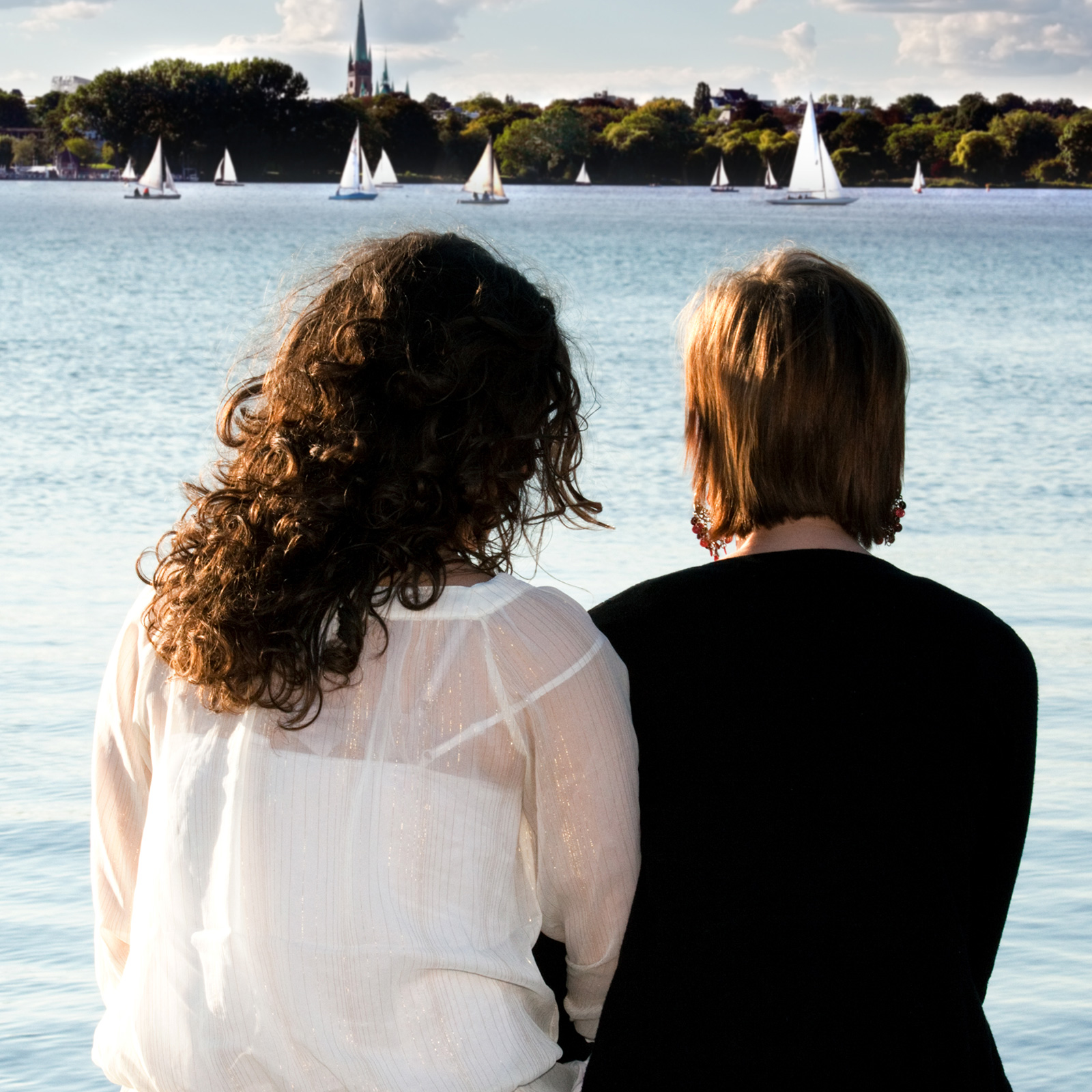 Two people sitting next to each other on pier watching sailboats.