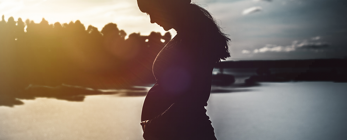 A pregnant woman silhouetted against a lake in sunset, looking down