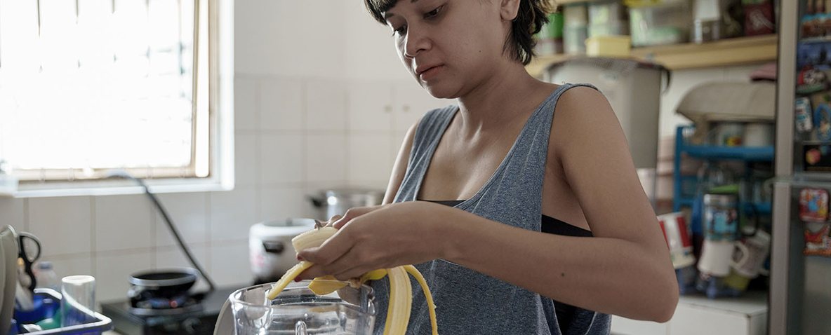 Pregnant woman in her kitchen, slicing a banana into a blender.