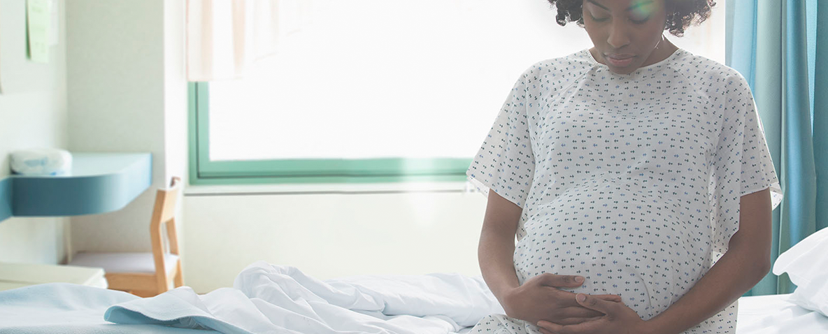 A pregnant woman in a hospital gown sitting on a hospital bed cradling her abdomen.
